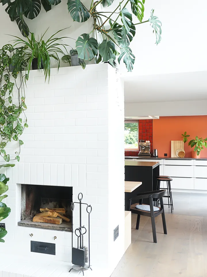 Wohnbereich mit weißem Kamin, Pflanzen und offener Küche im Hintergrund.