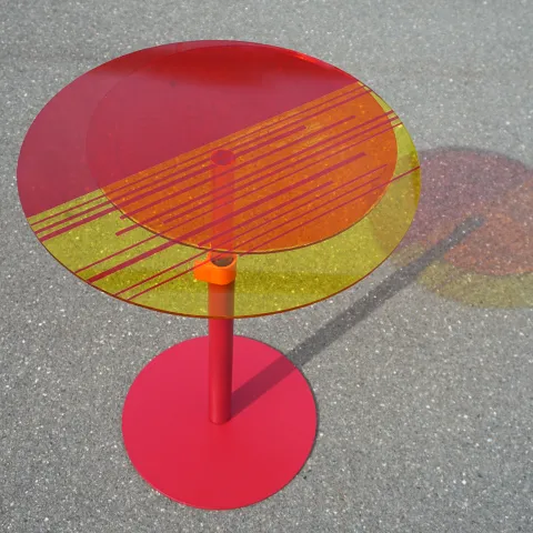 Runder Tisch mit farbiger, transparenter Tischplatte und rotem Fuß.
