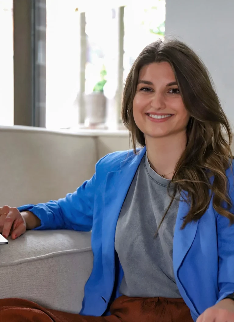 Porträt einer lächelnden Frau mit braunen Haaren, die auf einem Sofa sitzt. Sie trägt einen blauen Blazer und ein graues Oberteil, im Hintergrund ein helles Fenster mit Pflanze